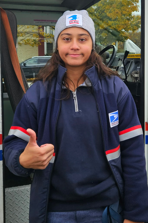 A woman in a postal uniform stands next to a postal delivery vehicle