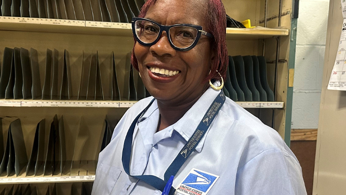 A woman in a blue USPS shirt stands in front of a letter sorting rack.