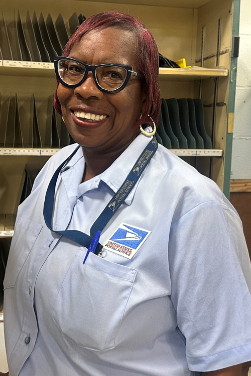 A woman in a blue USPS shirt stands in front of a letter sorting rack.