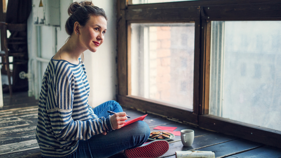 A woman sitting cross-legged on the floor with a pen in her right hand and paper on her knee