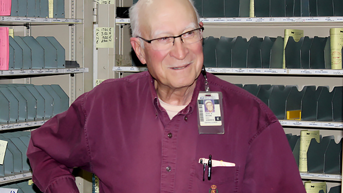 A man wearing glasses and a burgundy button-up shirt standing in front of a mail-sorting rack.