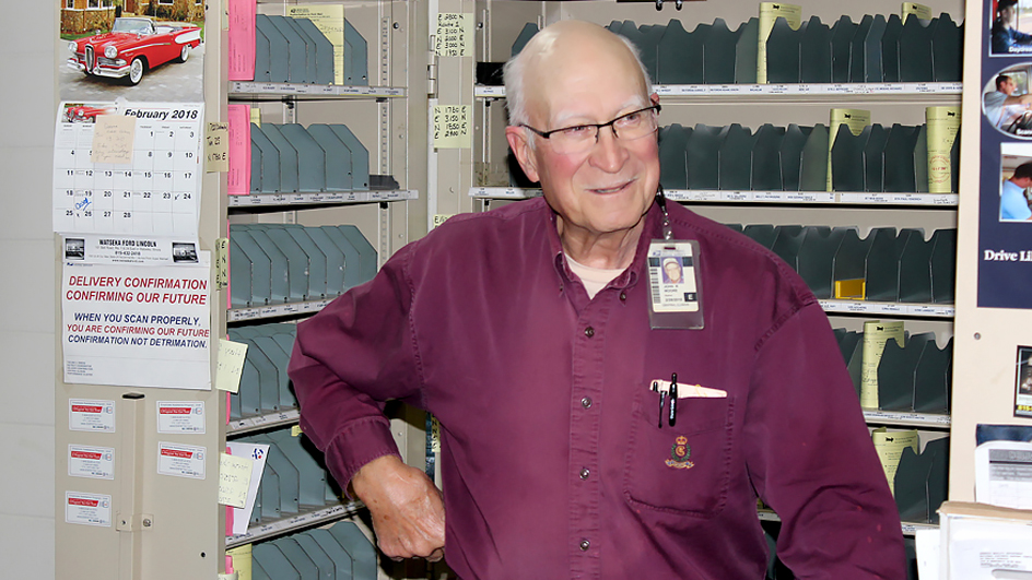 A man wearing glasses and a burgundy button-up shirt standing in front of a mail-sorting rack.