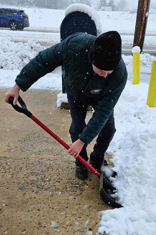 A man in a dark jacket shovels snow from a sidewalk.