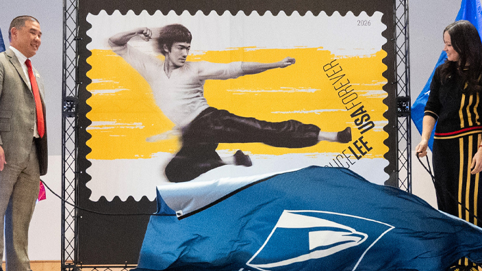 Three people stand on both sides of an enlarged image of the Bruce Lee stamp on stage