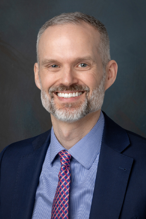 A smiling man with a beard wearing a blue and red tie and dark jacket