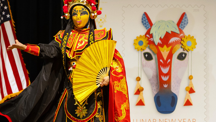 Performer wearing a decorative mask and Asian dress on stage with an enlarged image of the Year of the Horse stamp in the background