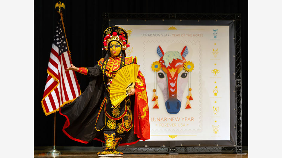 Performer wearing a decorative mask and Asian dress on stage with an enlarged image of the Year of the Horse stamp in the background