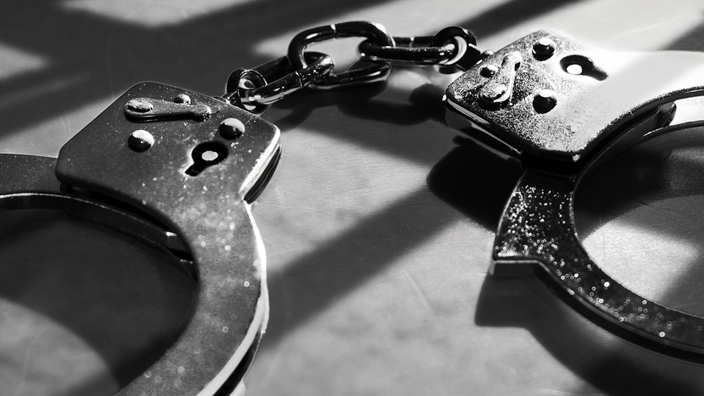 A conceptual photo of a pair of handcuffs on a floor, with light casting a shadow from jail cell bars nearby