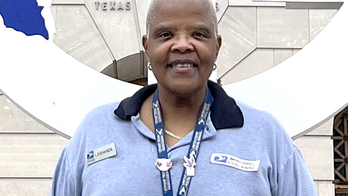 Female USPS employee wearing blue stands outside of a Post Office building.