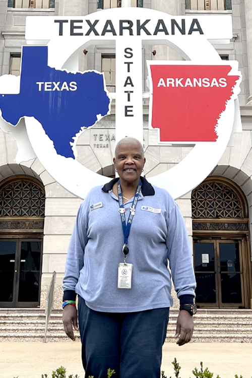 Female USPS employee wearing blue stands outside of a Post Office building.