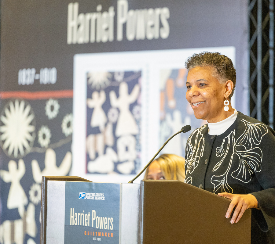A woman behind a podium with an enlarged image of the Harriet Powers stamps in the background