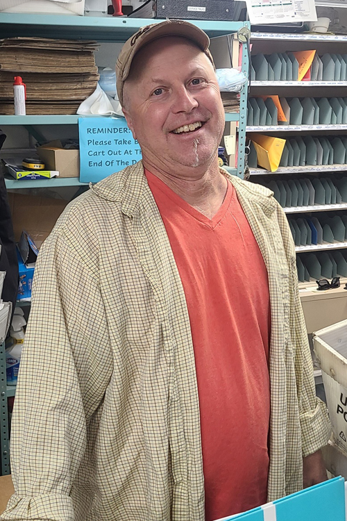 Man in baseball cap and orange shirt standing amongst bins of mail