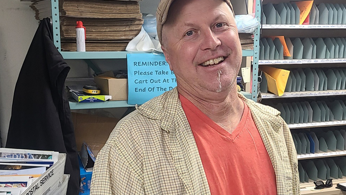 Man in baseball cap and orange shirt standing amongst bins of mail
