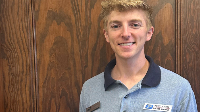 A younger man wearing a blue USPS shirt holds a white USPS mail container