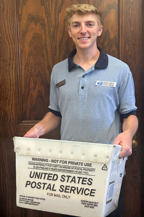 A younger man wearing a blue USPS shirt holds a white USPS mail container