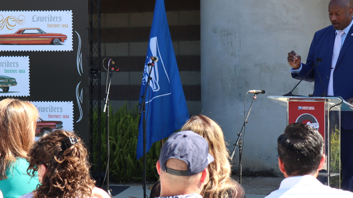 Man in blue suit standing behind a podium and to the right of enlarged images of USPS Lowriders stamps