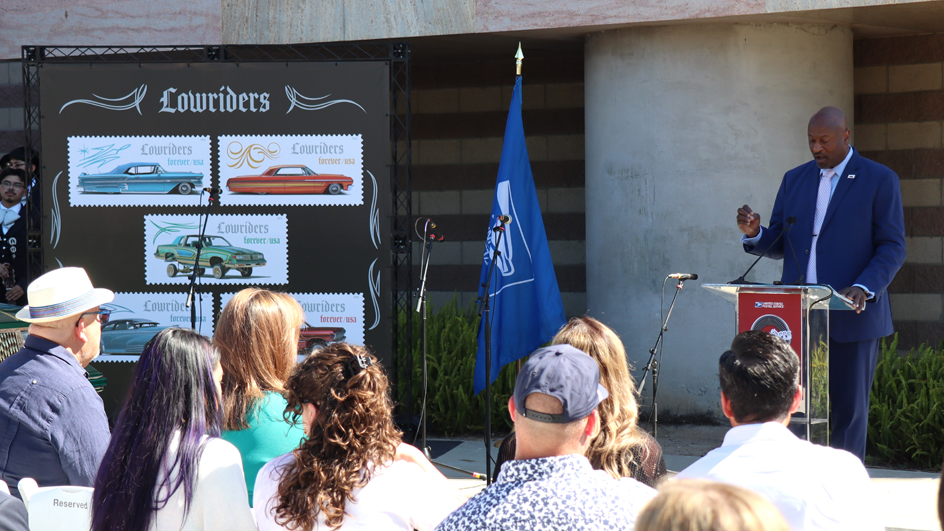 Man in blue suit standing behind a podium and to the right of enlarged images of USPS Lowriders stamps