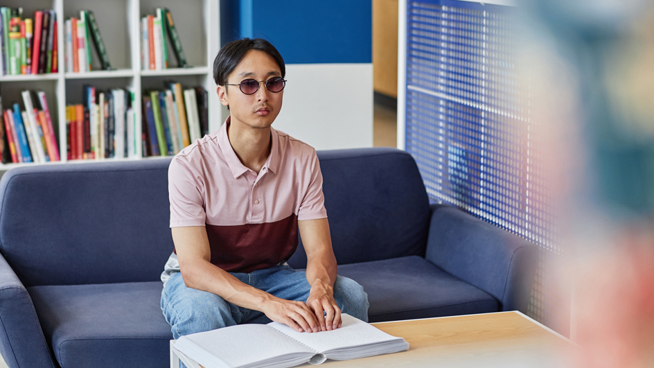 A man wearing dark glasses reading a book in tactile braille