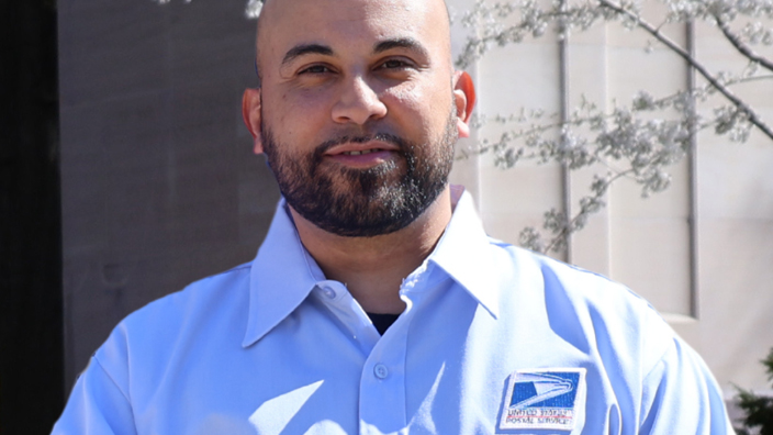 Postal worker in uniform standing outdoors near blooming trees and a park bench
