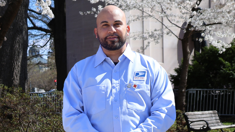 Postal worker in uniform standing outdoors near blooming trees and a park bench