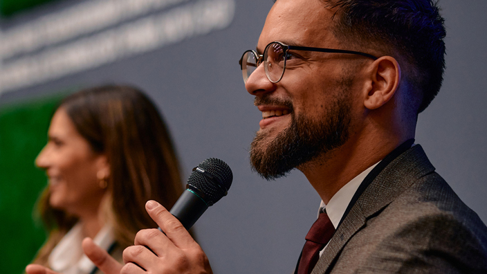 A man in a suit sitting in a chair and holding a microphone and two women sitting beside him in the background