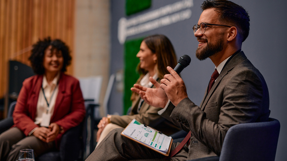 A man in a suit sitting in a chair and holding a microphone and two women sitting beside him in the background