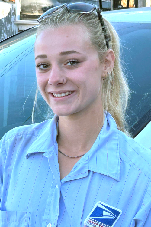 A woman wearing a blue Postal Service button-up shirt and standing in front of a vehicle