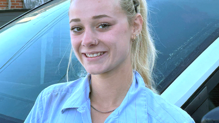 A woman wearing a blue Postal Service button-up shirt and standing in front of a vehicle