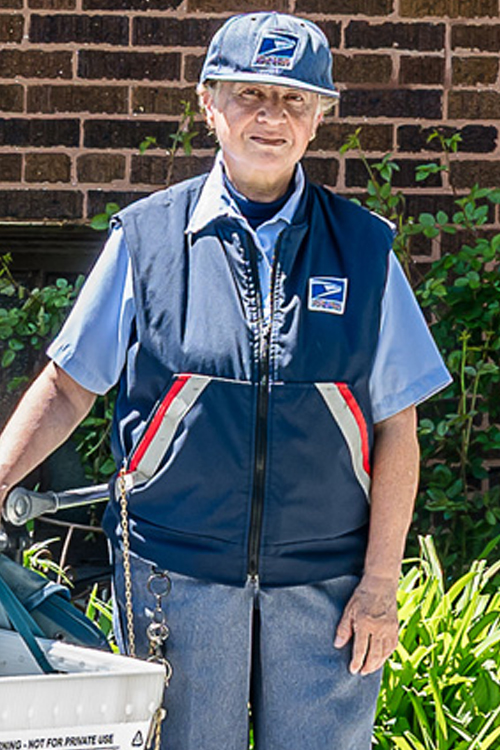 A USPS employee in uniform standing on a sidewalk and next to a mail cart