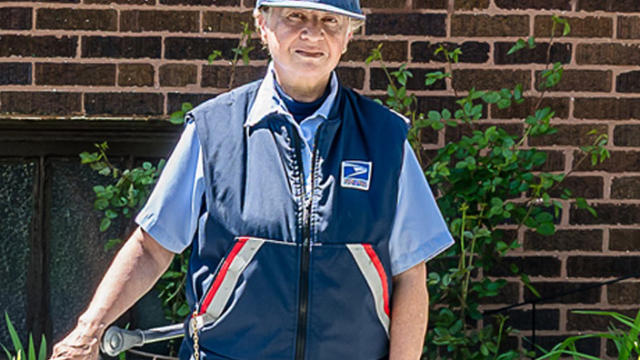 A USPS employee in uniform standing on a sidewalk and next to a mail cart