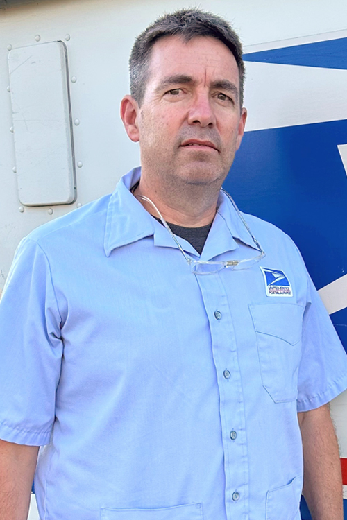 USPS employee in a blue button-up shirt standing in front of a delivery vehicle