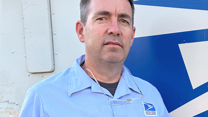 USPS employee in a blue button-up shirt standing in front of a delivery vehicle