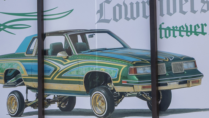 Large images of Lowrider stamps on a green and blue backgrounds filling up two large windows of a building.
