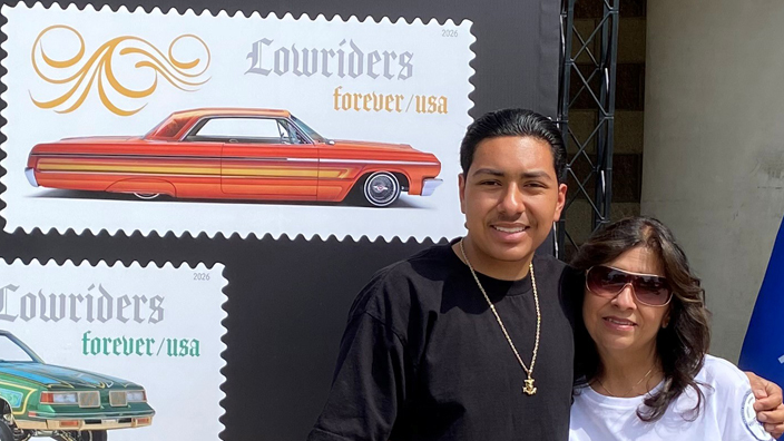 Three people standing outdoors by Lowriders stamp display next to a postal flag
