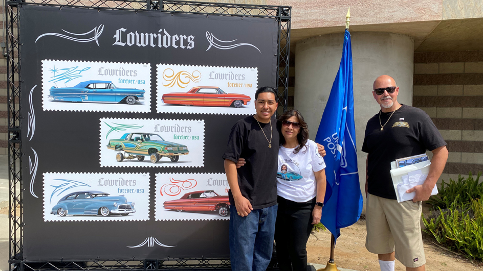 Three people standing outdoors by Lowriders stamp display next to a postal flag