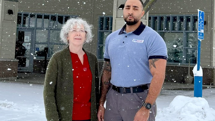 Two USPS employees standing in a snow-covered parking lot in front of a Post Office building