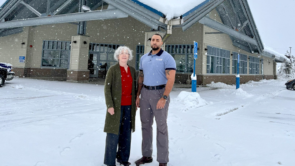 Two USPS employees standing in a snow-covered parking lot in front of a Post Office building