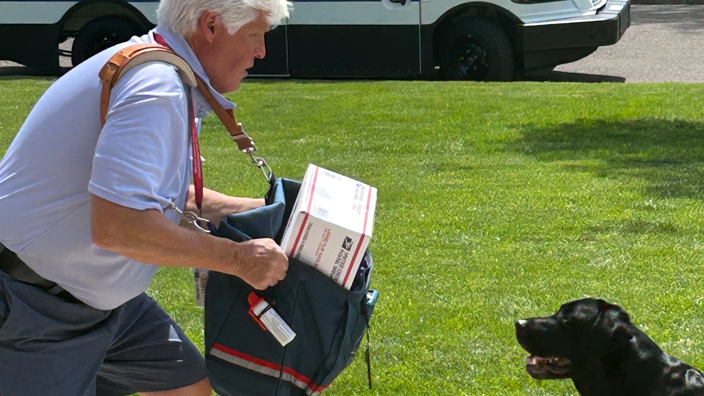 USPS letter carrier in a front yard holding up a satchel between himself and a black dog