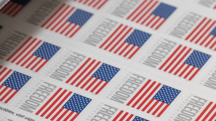 A sheet of stamps bearing the U.S. flag and the word “Freedom” rolls off a printing press.