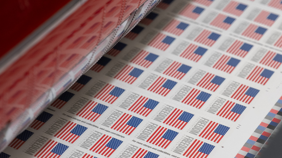 A sheet of stamps bearing the U.S. flag and the word “Freedom” rolls off a printing press.