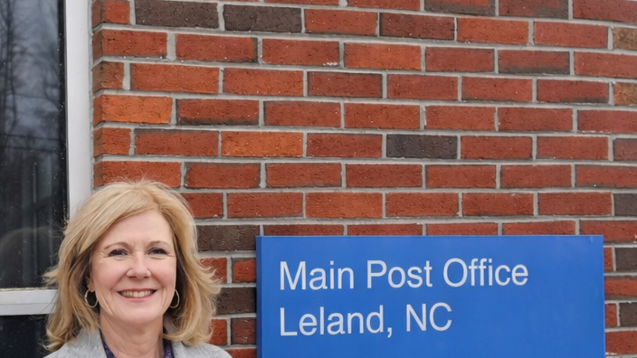 A woman wearing a gray coat and standing outside of the Leland, NC, Post Office building