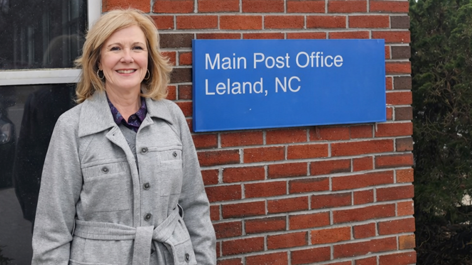 A woman wearing a gray coat and standing outside of the Leland, NC, Post Office building