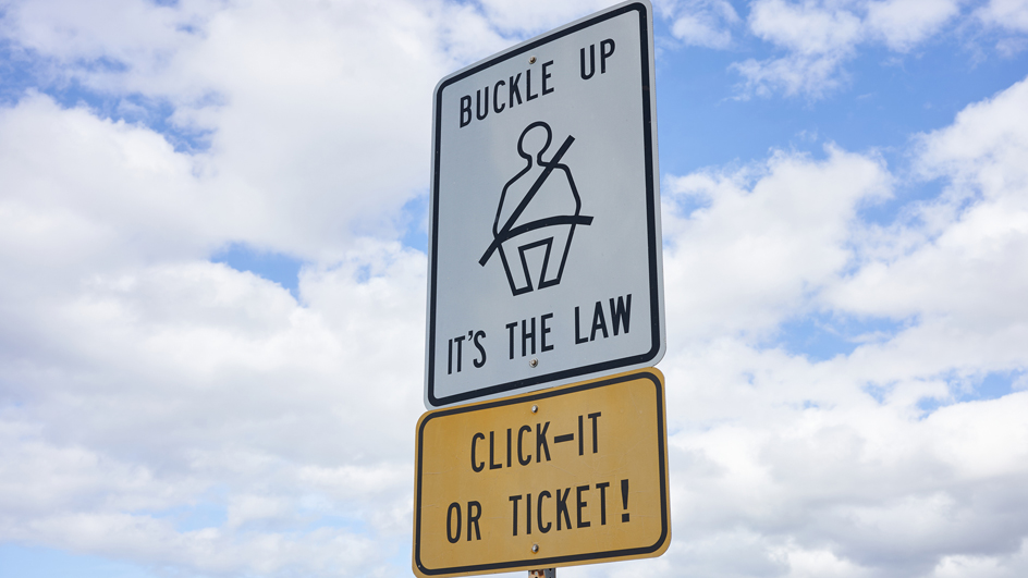 Buckle up sign and 'Click It or Ticket' warning sign against blue sky and clouds