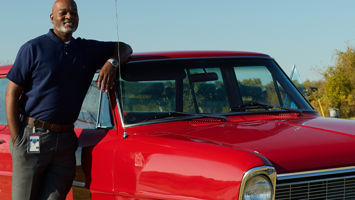 A man wearing dark slacks and a blue polo shirt leaning against a red car
