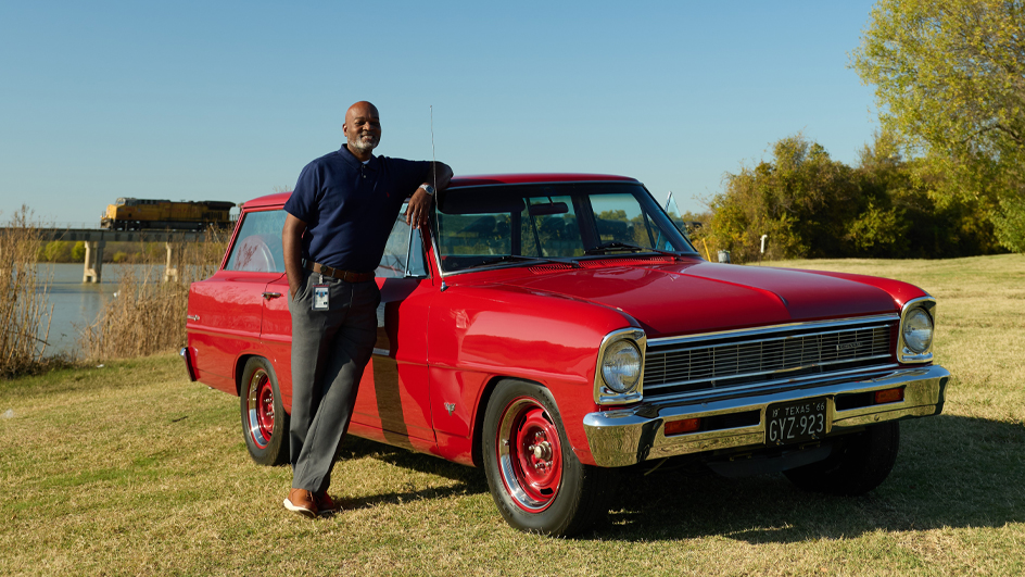 A man wearing dark slacks and a blue polo shirt leaning against a red car