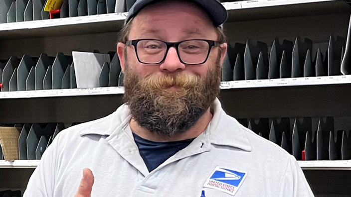 USPS employee giving a thumbs-up and wearing a light-colored polo shirt and blue hat