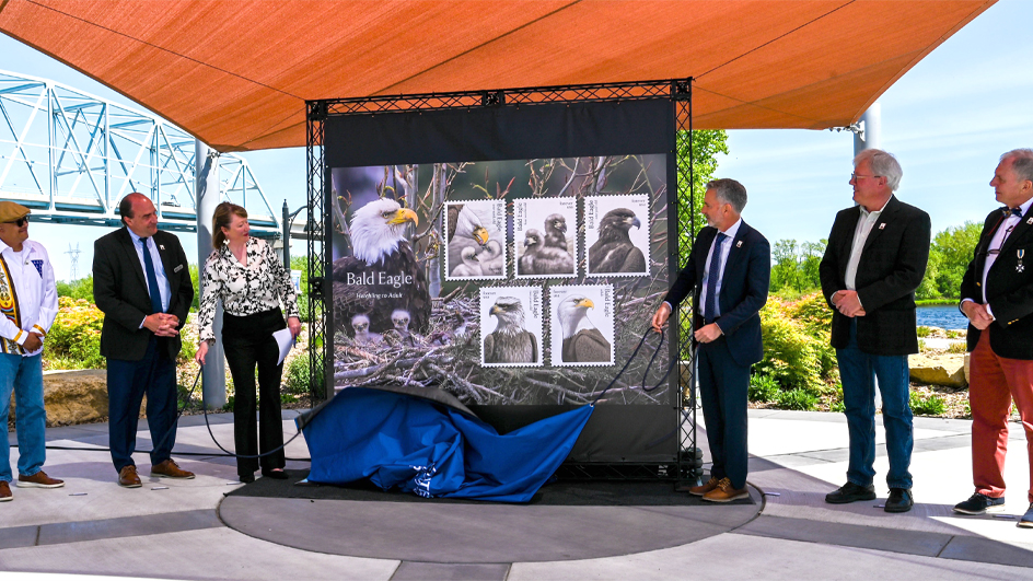 Three people standing outdoors on either side of an enlarged image of the Bald Eagle: Hatchling to Adult stamps