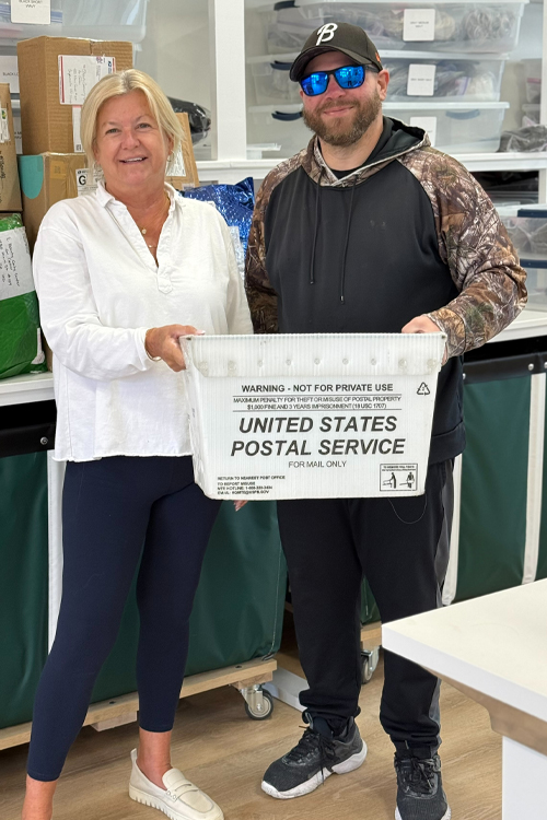 A woman in a white blouse and blue pants standing next to a man wearing black, both holding up a USPS mail container