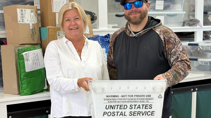 A woman in a white blouse and blue pants standing next to a man wearing black, both holding up a USPS mail container