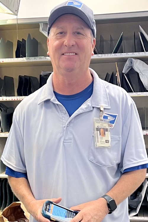 USPS employee wearing blue shorts, a blue shirt and a blue baseball cap standing in front of a sorting station